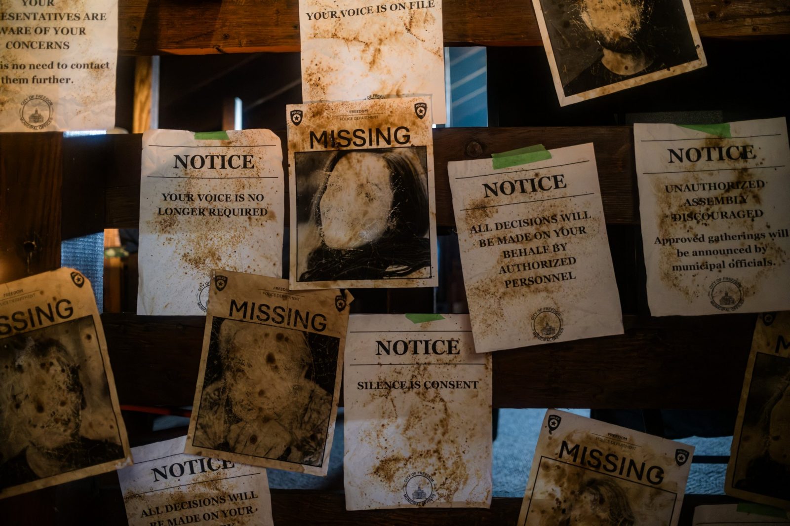 Weathered notices and missing posters on a barn wall, reading "Your voice is no longer required" and "Silence is consent"