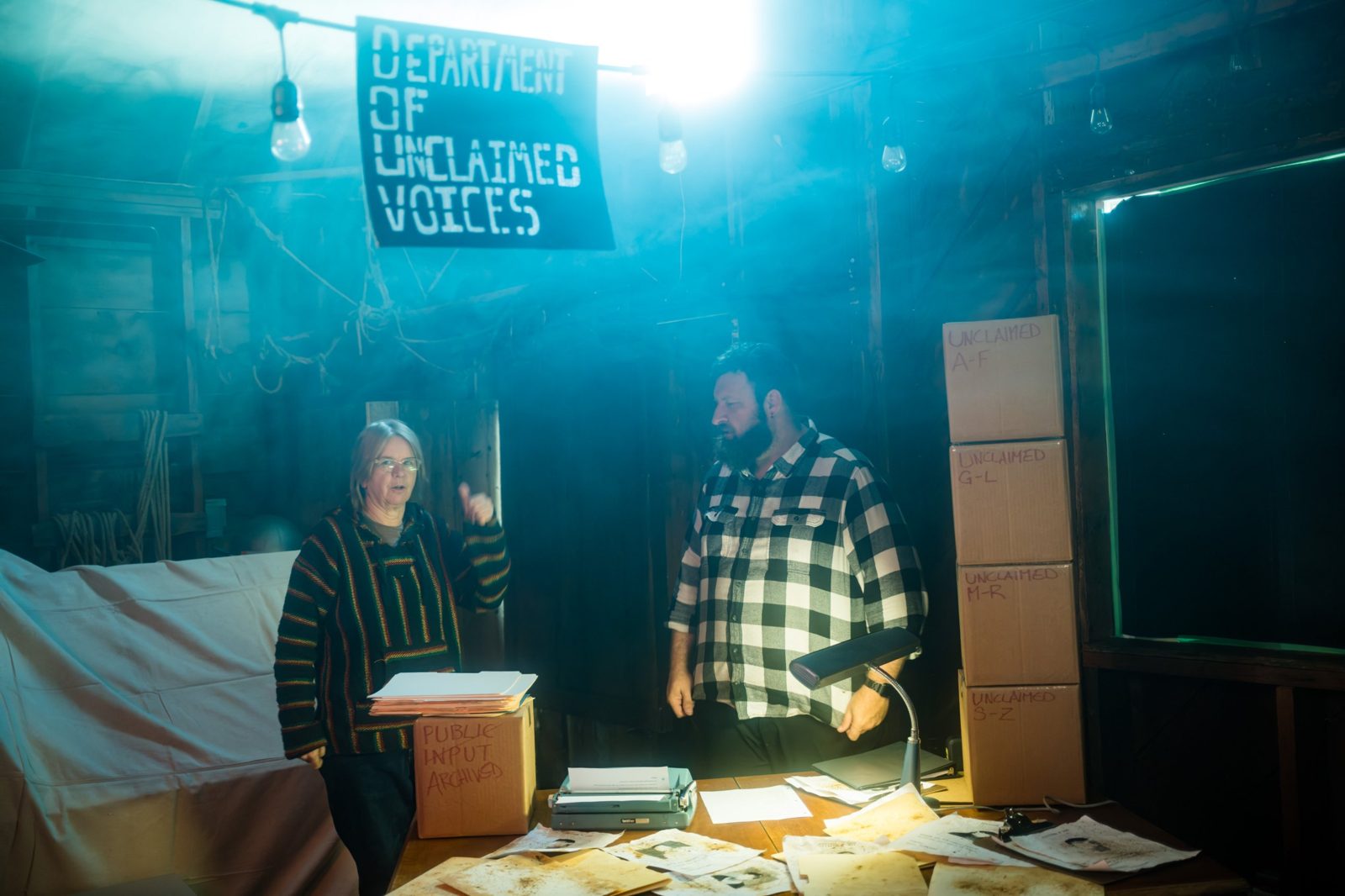 A desk under a sign reading "Department of Unclaimed Voices," with two actors and boxes labeled "Unclaimed A-F" and "Public input archived"