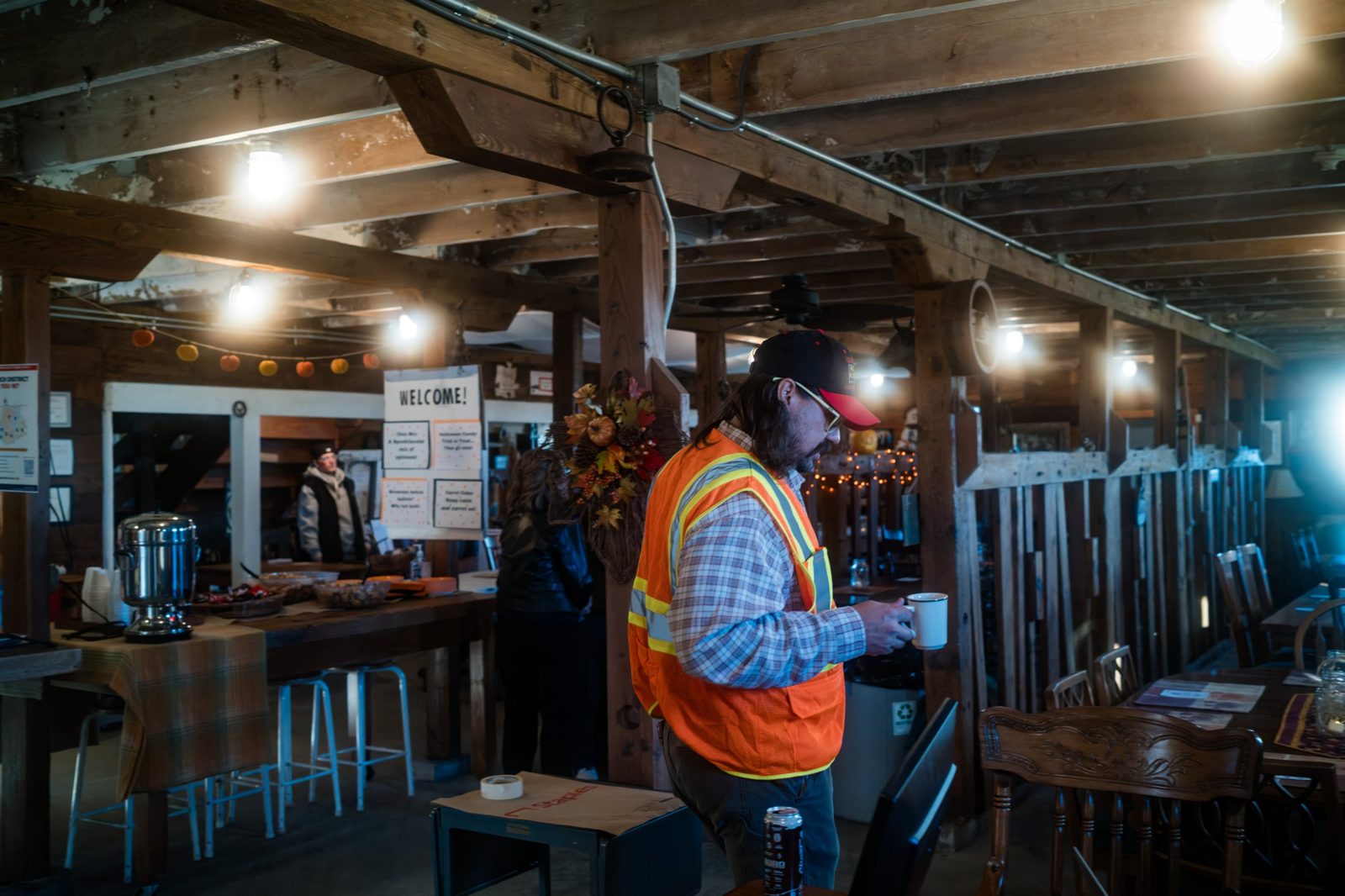 Organizer Jordan Deffenbaugh in an orange safety vest, holding a coffee mug in the barn's gathering space, with a "Which district are you in?" poster on the wall