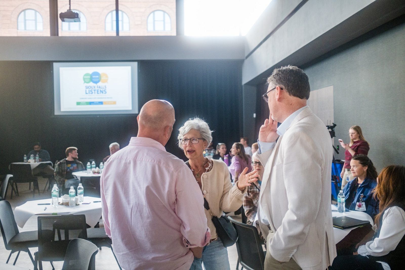 Neighbors talk between rounds, with the Sioux Falls Listens screen in the background